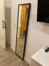 Espejo de cuerpo entero apoyado en la pared blanca de la habitación, con el televisor montado y la puerta abierta